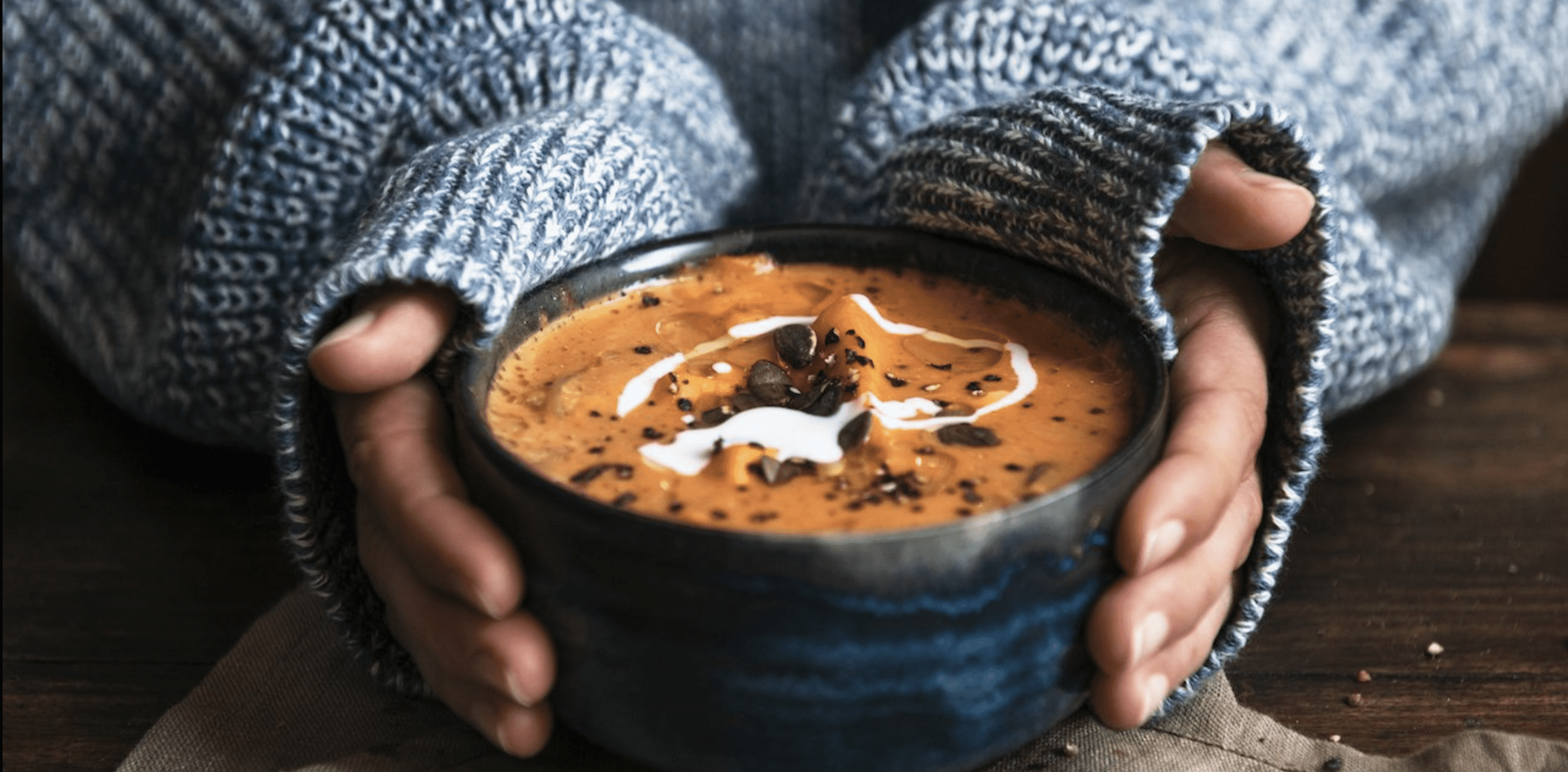 A woman wearing a cozy, blue sweater holds a bowl of soup